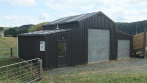 Rural shed with a gabled roof and durable black steel cladding, featuring roller doors and a side entry door, set in a pastoral landscape with hills and trees in the background.