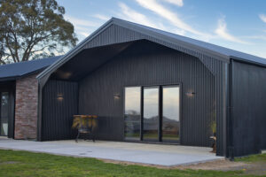 Modern black corrugated steel building with a gabled roof, large sliding glass doors, and a concrete patio, set against a backdrop of trees and a blue sky.