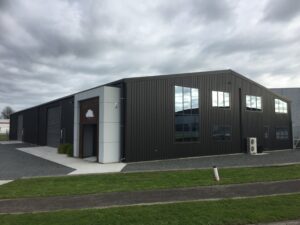 Modern industrial building with sleek black corrugated steel cladding, featuring large reflective windows, multiple roller doors, and a distinct white panel entrance with a tree logo. Surrounded by well-maintained gravel pathways and lush green lawns, this versatile commercial structure stands out against a dramatic cloudy sky, ideal for warehouse, office, or multi-purpose use