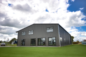 Modern black steel-frame barndominium with large glass windows and doors, set on a lush green lawn under a partly cloudy sky.